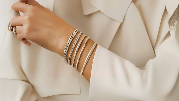 Elegant wrist adorned with four different silver bracelets—curb chain, beaded, cuff, and flat styles paired with a soft cream-colored outfit.