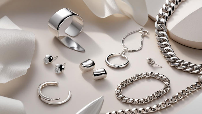 Flat lay of polished silver jewelry including chunky silver chains, hoops, bracelets, and rings on soft satin backdrop.